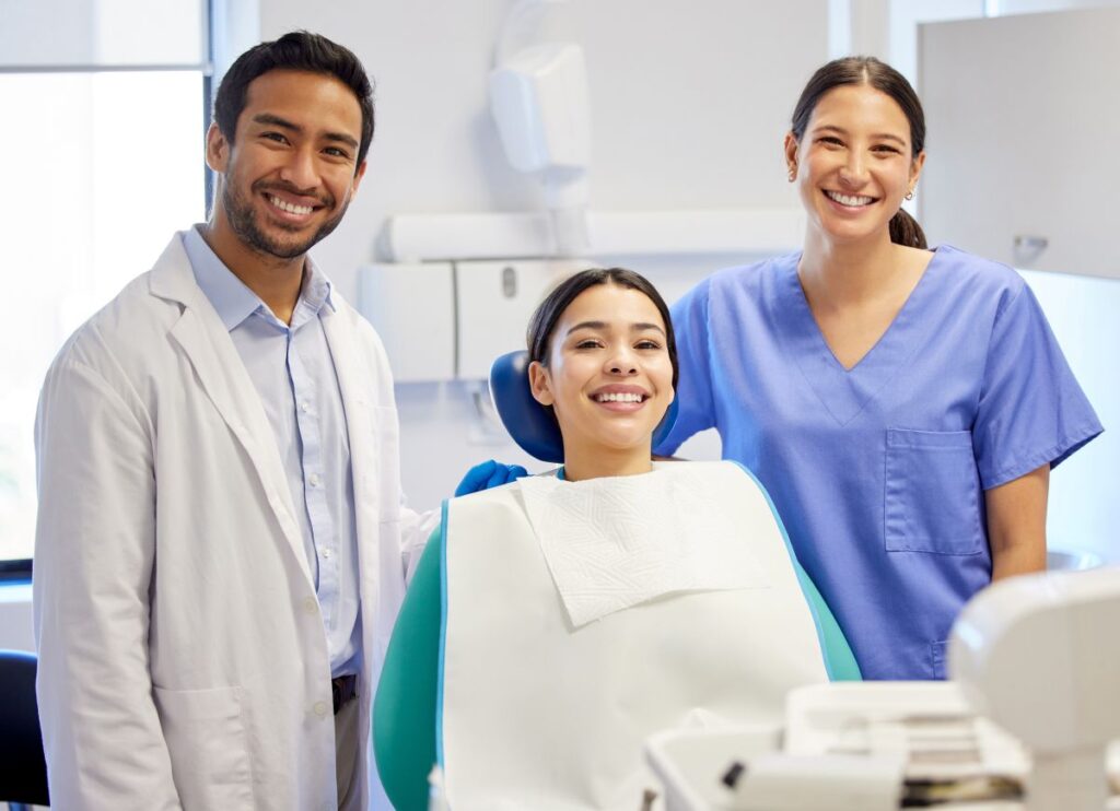 Smiling patient in a dental chair with a dentist in a white coat and a dental assistant in blue scrubs beside her in a clinical room.