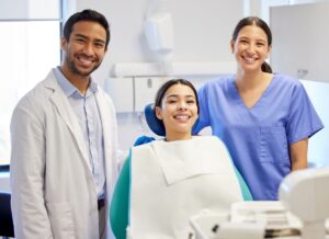 portrait-of-a-young-woman-having-a-consultation-wi-2026-01-09-09-55-09-utc - Serenity Healthy Smiles Smiling patient in a dental chair with a dentist in a white coat and a dental assistant in blue scrubs beside her in a clinical room.