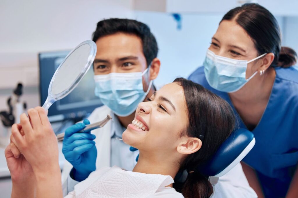 Smiling patient in a dental chair looking at a mirror as a dentist and assistant in masks prepare a checkup.